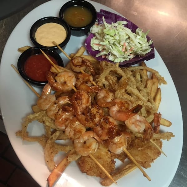 Shrimp skewers, french fries, onion rings, coleslaw, and three dipping sauces on a white plate.